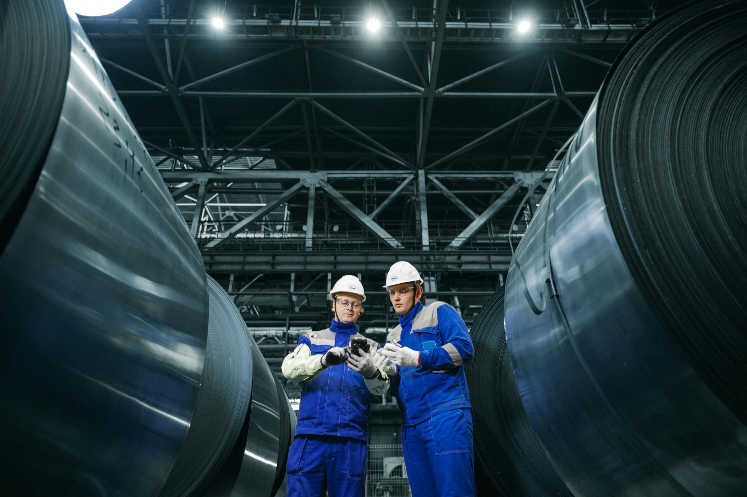 Two workers in protective gear examine steel coils in a modern industrial facility.